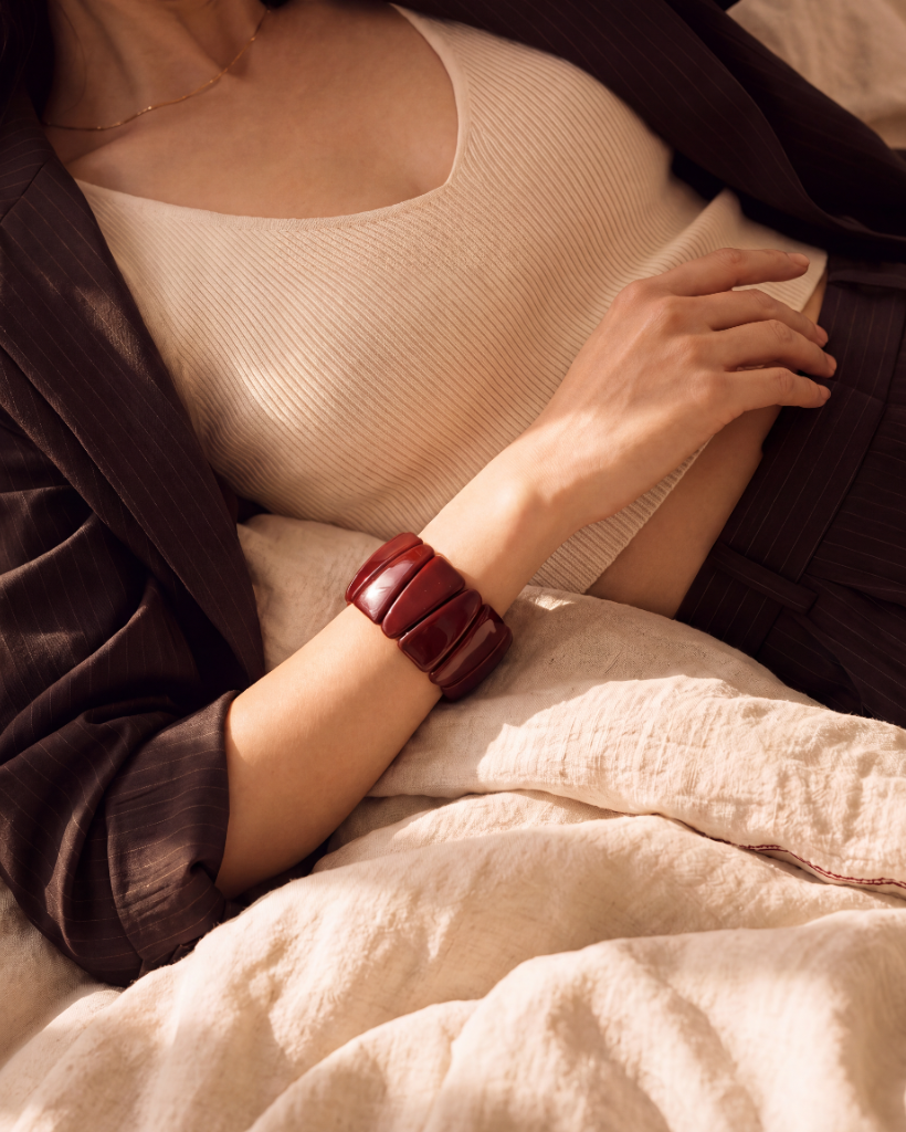 A close-up lifestyle shot of a model wearing a chunky, segmented dark red leather bracelet on her wrist, styled with a cream ribbed tank top, a dark brown blazer, and a matching textured blanket.