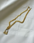 A chunky gold Cuban link chain necklace with a small gold handbag pendant, laid out on a white satin fabric background.