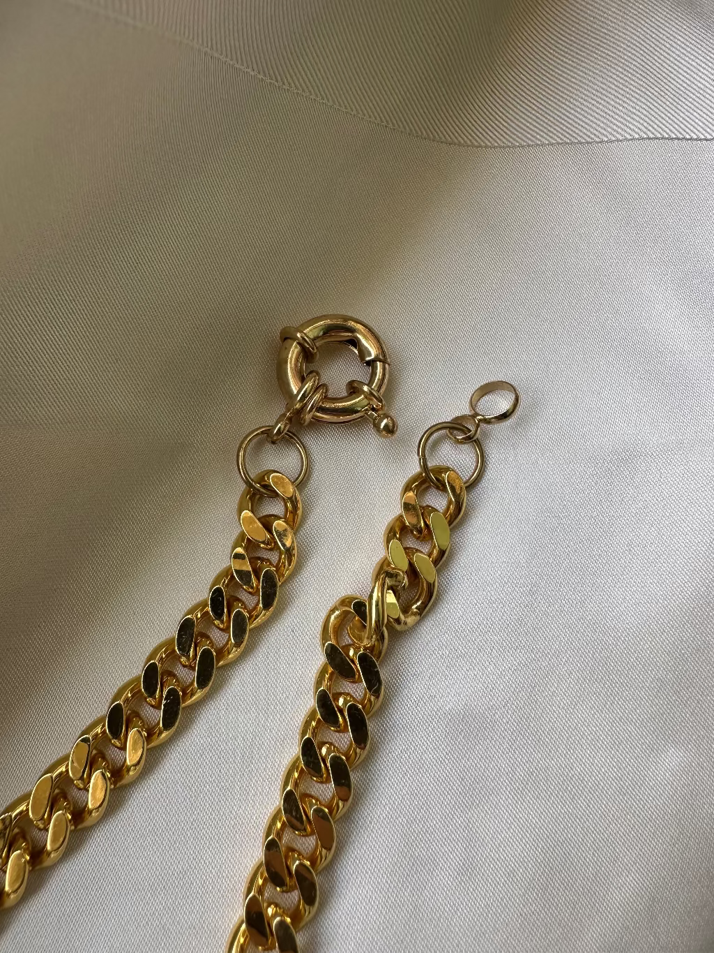 A close-up of a gold-toned chain strap with a lobster clasp and a small ring, laid on a white fabric background.