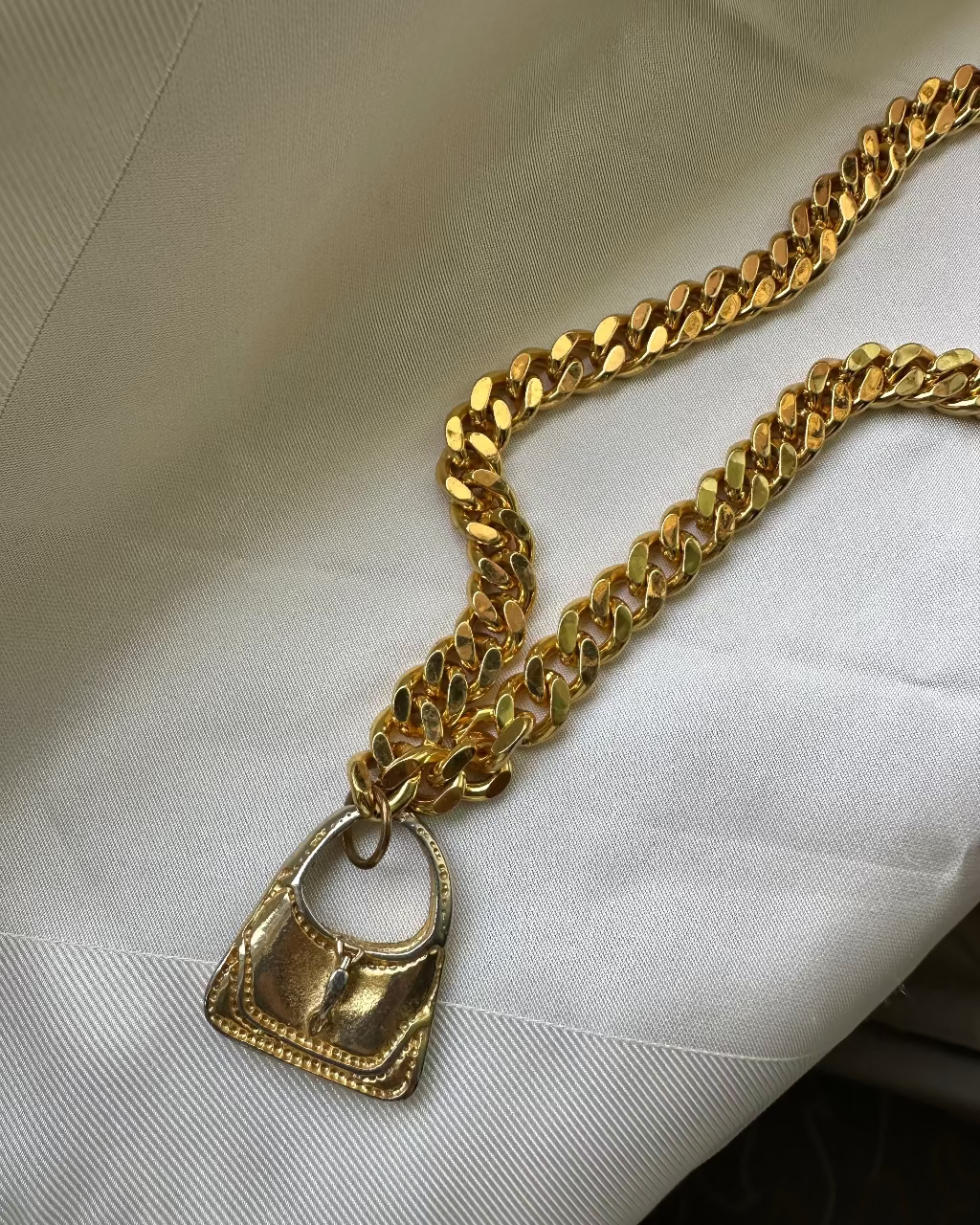 A chunky gold-tone Cuban link chain necklace with a small gold handbag pendant, displayed on a white textured fabric background.