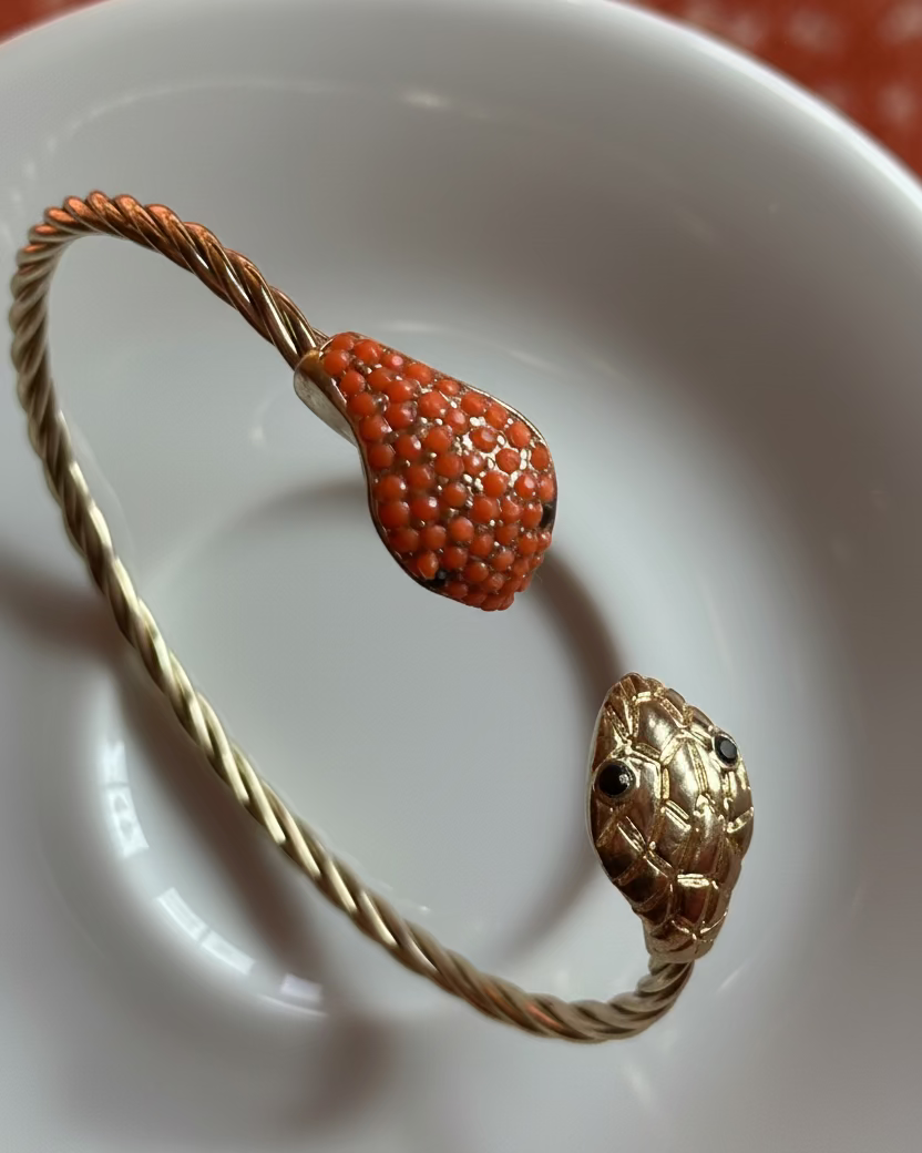 A gold-toned twisted wire cuff bracelet with a teardrop-shaped orange coral inlay and a gold snake head charm with small black eyes, held by a hand over a white ceramic dish.