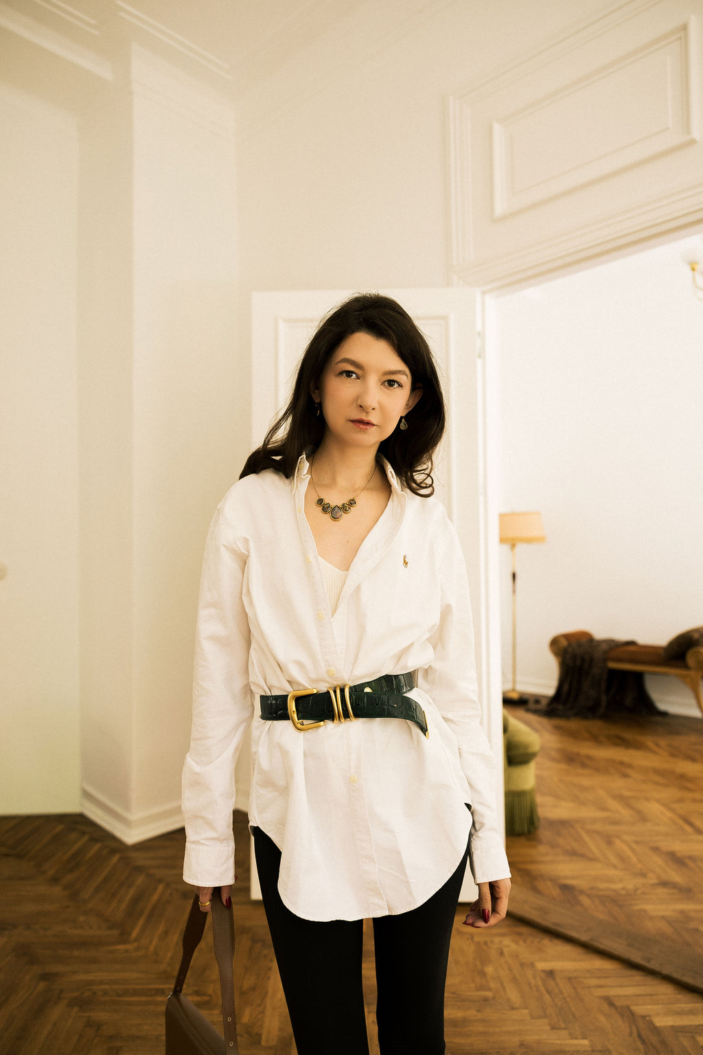 A woman stands indoors wearing a white long-sleeve button-up shirt with a dark green belt featuring a gold buckle, paired with black pants. She is holding a brown leather bag in a bright room with cream walls and wood parquet flooring.