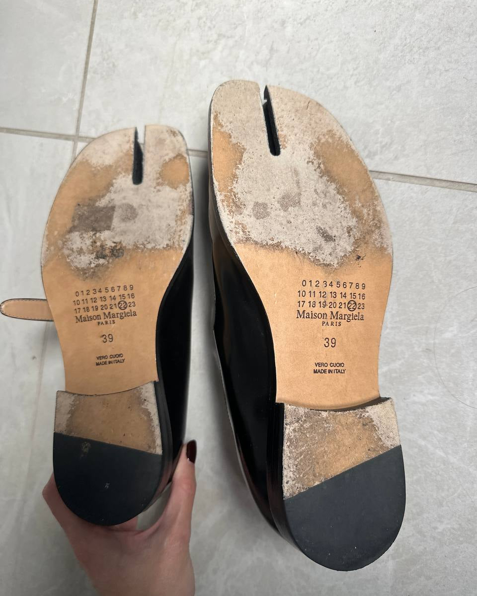 A pair of black leather tabi-style shoes shown from the sole, held by a hand over a light gray tiled floor. The tan insoles display the brand name 'Maison Margiela' and size '39'.