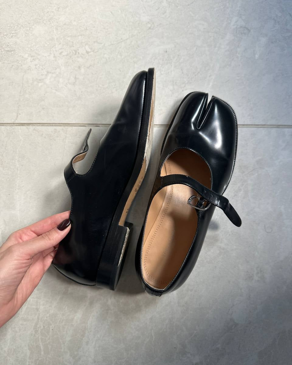 A pair of glossy black Mary Jane shoes with a strap and buckle, shown on a gray tiled floor. A hand is holding one shoe to display its side profile.