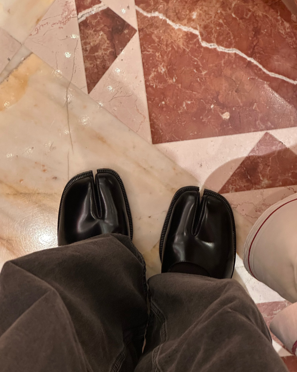 A person wearing black leather tabi-style shoes and dark brown pants, standing on a polished beige and reddish-brown marble floor.