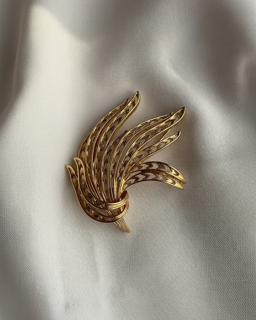 A gold-toned feather brooch with delicate cutout detailing and small dark inlay accents, displayed on a soft white fabric background.