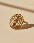A small oval gold-tone decorative button or charm with an ornate monogram design, shown on a plain light gray background.