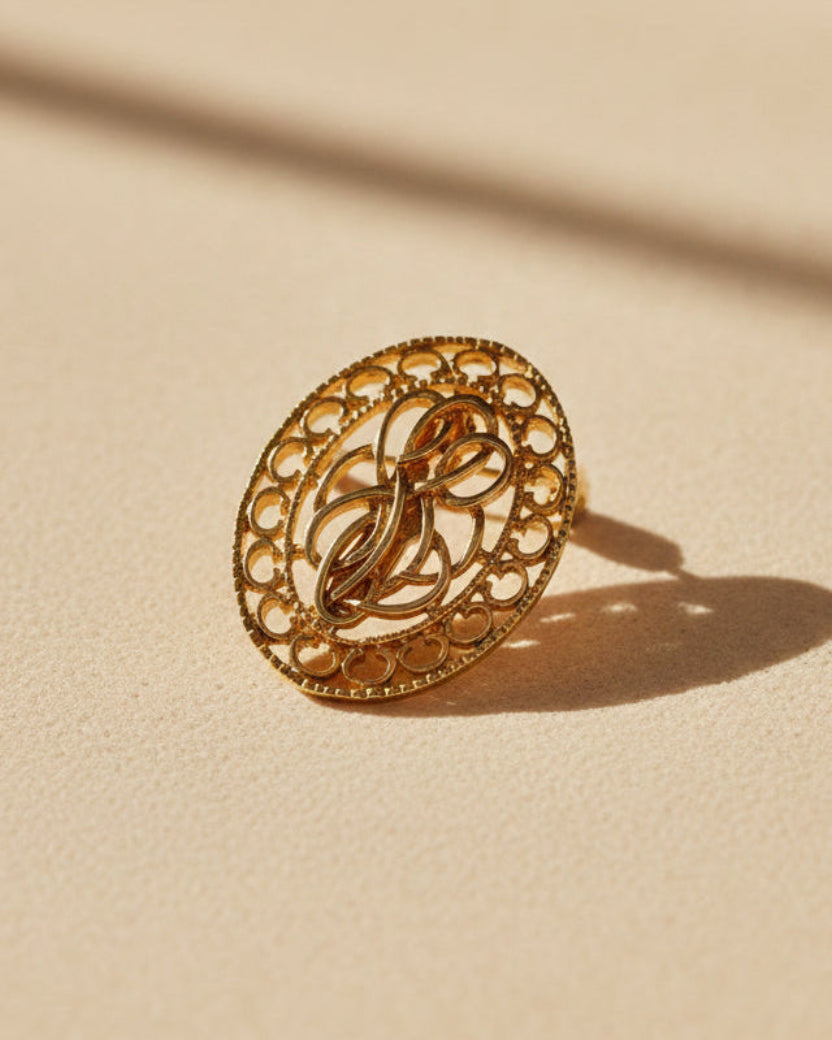 A small oval gold-tone decorative button or charm with an ornate monogram design, shown on a plain light gray background.