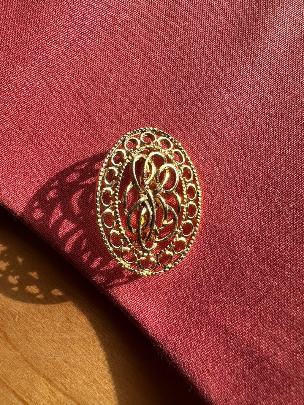 An oval gold filigree brooch with an interlocking monogram design, resting on a textured dark red fabric surface with a wooden surface visible in the lower-left corner.