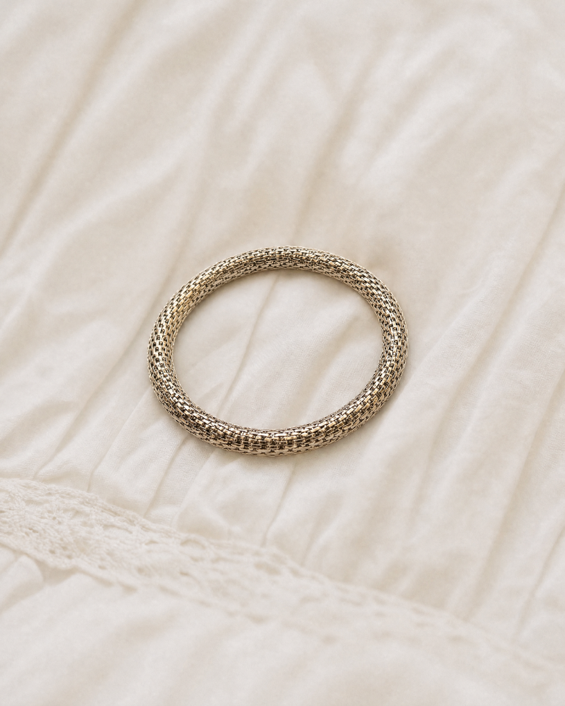 A textured metallic silver bangle bracelet resting on a soft, off-white pleated fabric background.