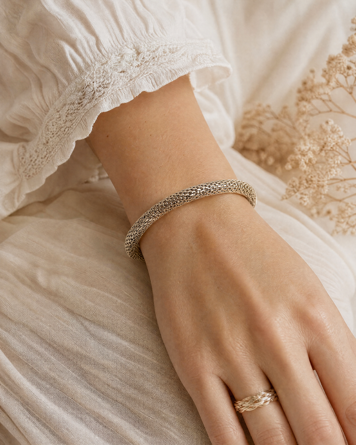 A close-up of a person's hand and wrist wearing a textured silver bangle bracelet, styled with a matching textured gold ring on the finger, set against a soft cream-colored dress and dried flowers.