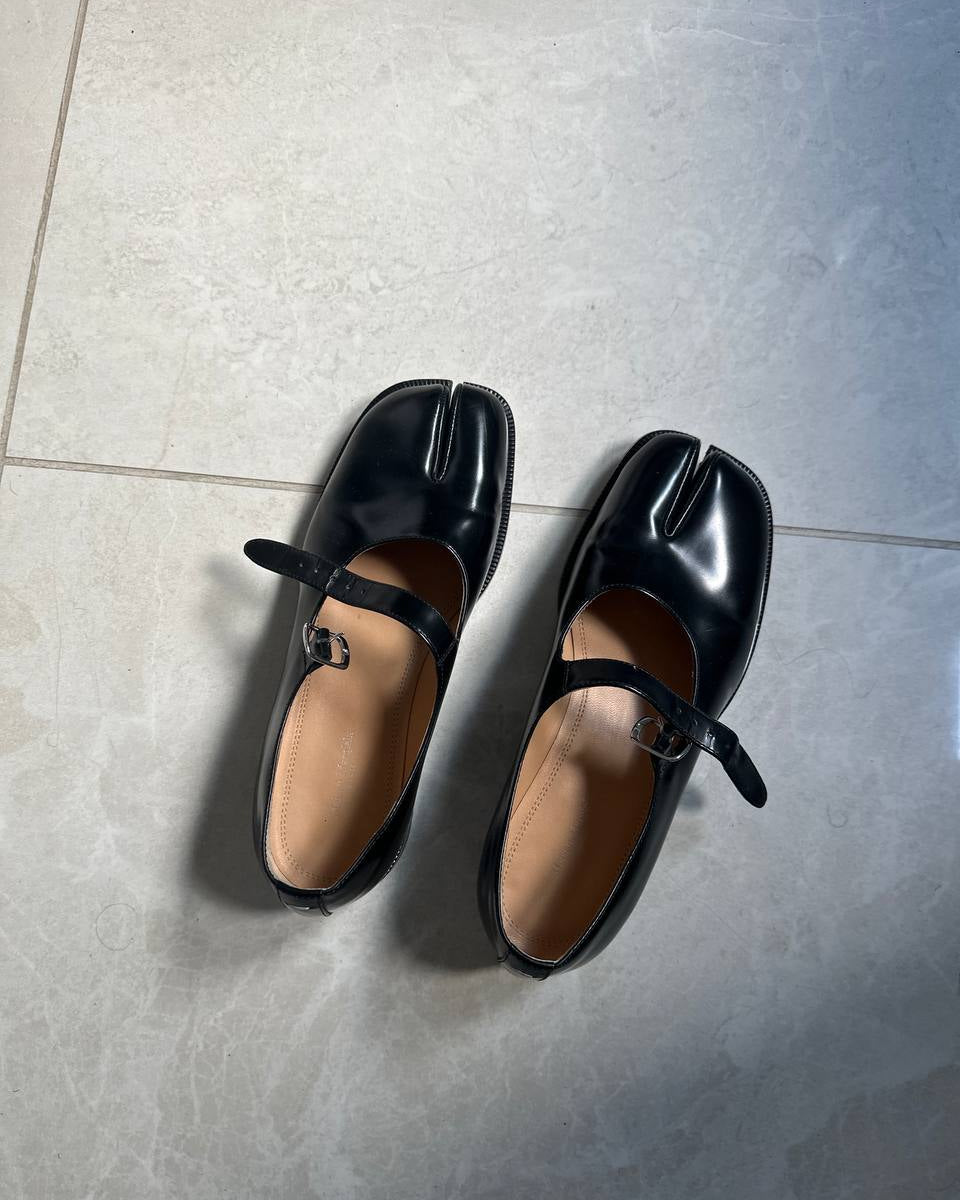 A pair of glossy black leather tabi-style Mary Jane shoes with ankle straps, displayed on a light gray tiled floor.