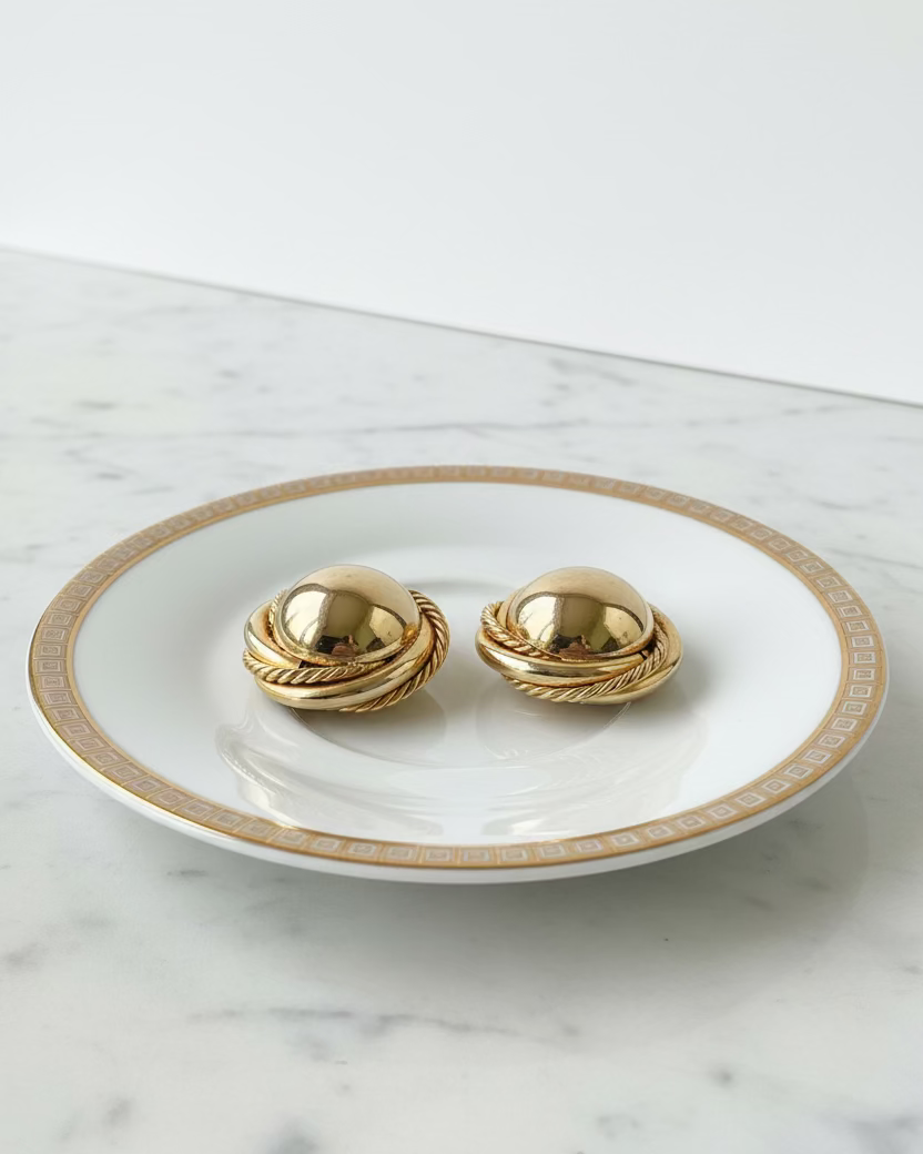 Two gold-toned oval earrings with twisted rope detailing and a central metal connector, displayed on a white ceramic plate with a gold geometric border.