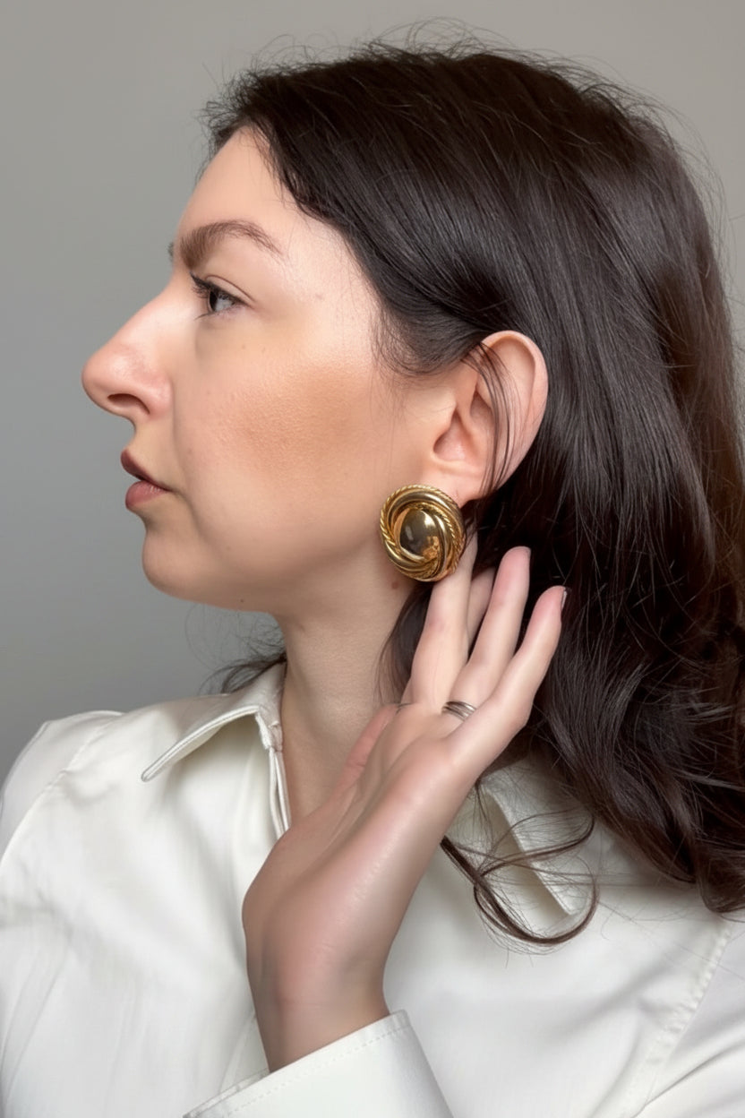 A woman in profile wearing a large, circular gold earring with a textured spiral or swirl design, paired with a white collared blouse.
