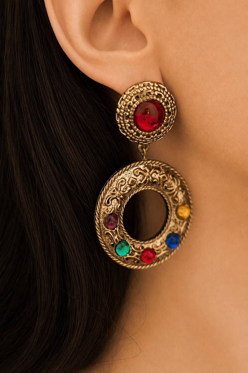 A close-up of a model's ear wearing a large gold-toned drop earring with a red gemstone at the top and a circular filigree charm decorated with small multicolored gemstones.