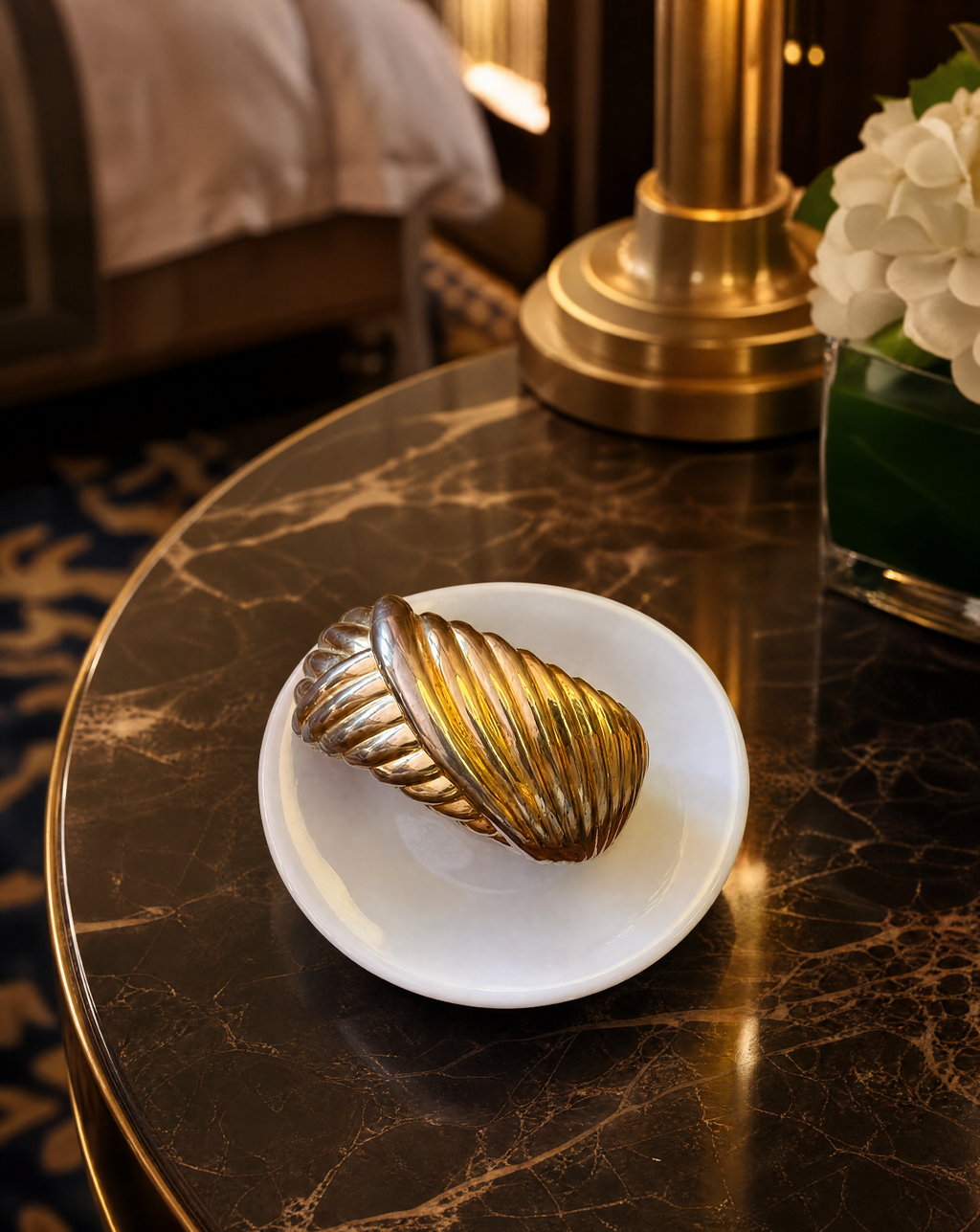 A metallic gold ribbed decorative object resting on a small white plate, placed on a dark marble table in an elegant interior setting with a lamp base and flowers visible in the background.