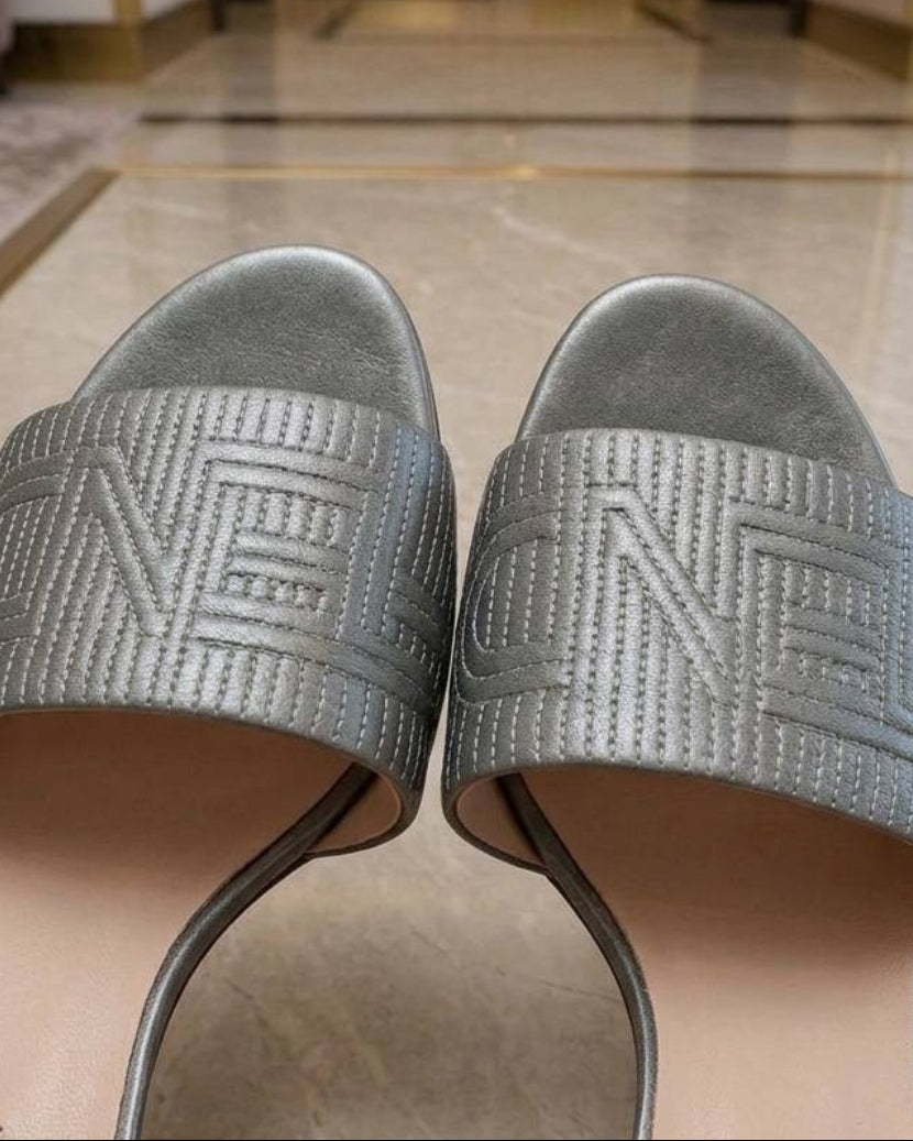 A pair of Fendi slide sandals with metallic silver straps featuring the embossed FF logo, resting on a marble table in a luxury interior.