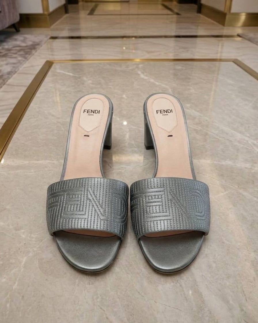 A pair of Fendi metallic silver slide sandals with embossed Greek key patterns, displayed on a polished marble floor in an elegant interior setting.